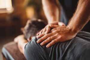 Patient discussing back pain with a clinician beside an examination table and anatomy chart.